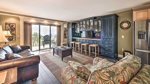 A spacious living room with a dark leather sofa, a floral-patterned couch, and a wooden coffee table. A kitchen with dark blue cabinets and a stainless steel refrigerator is visible in the background. - PCB Condo w/ 2 Outdoor Pools & Beach Access! (Panama City)
