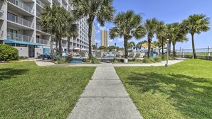 A hotel with a central pathway flanked by palm trees, a pool area, and a building with balconies. - PCB Condo w/ 2 Outdoor Pools & Beach Access! (Panama City)