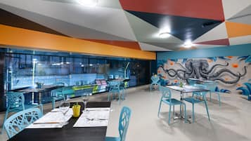 A modern restaurant with a vibrant mural, teal chairs, and a glass partition separating the dining area from the kitchen.