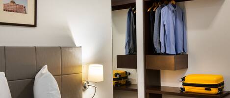 Habitación de hotel con cama, mesita de noche con lámpara, armario con ropa colgada, maleta y un cuadro en la pared.