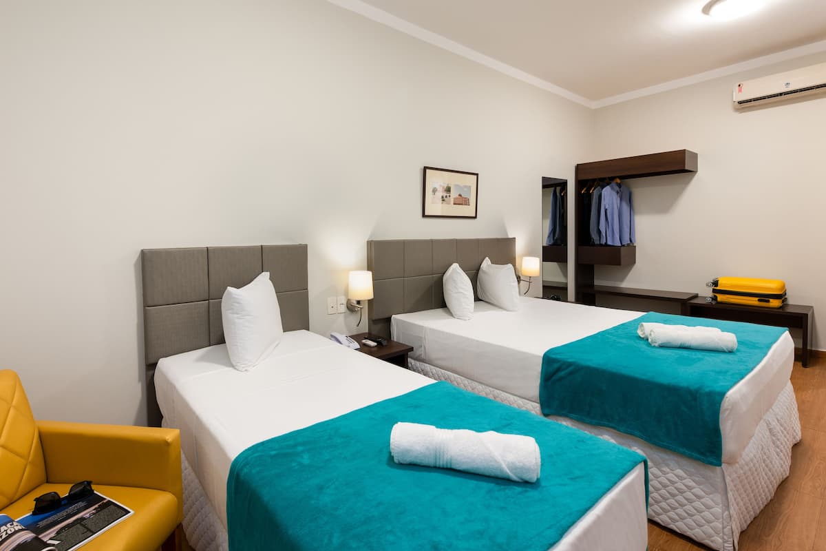 A hotel room with two beds, a chair, a small table, a wardrobe, and an air conditioning unit.