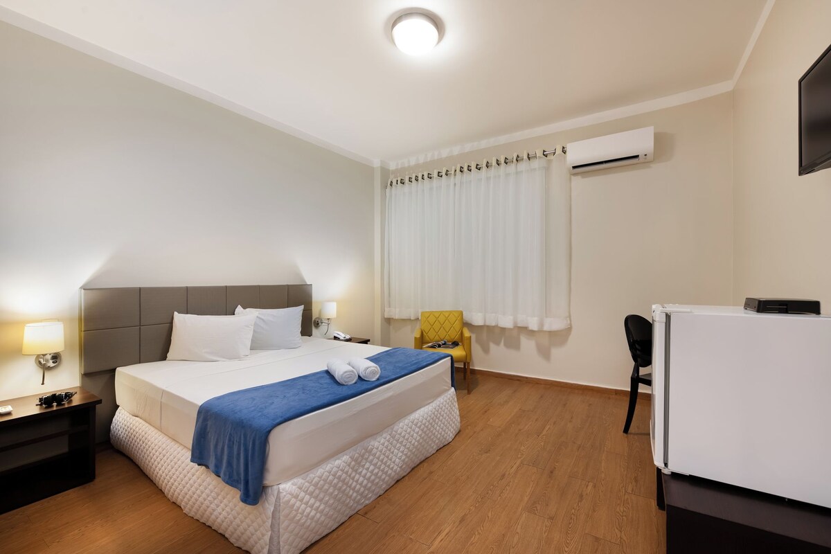 A hotel room with a bed, a desk, a chair, an air conditioning unit, and a television.