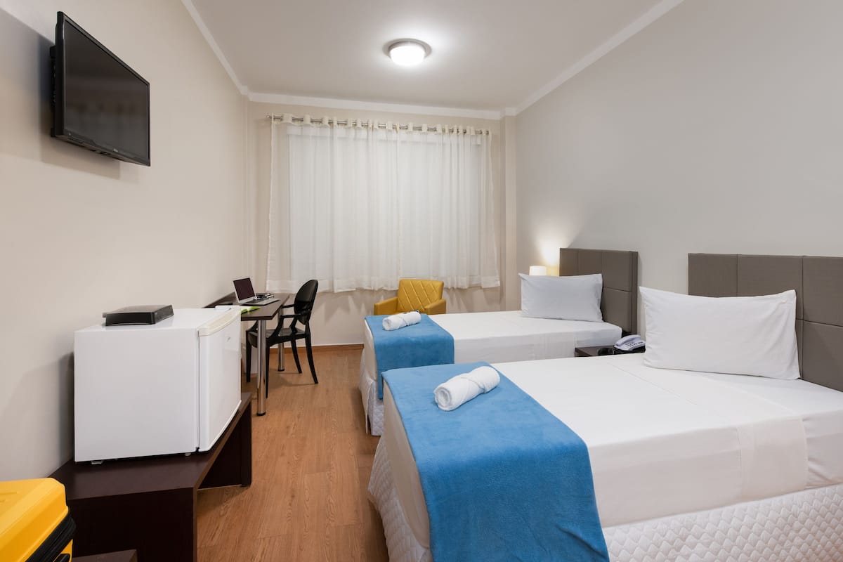 A hotel room with two beds, a desk, a chair, a TV, and a window with curtains.