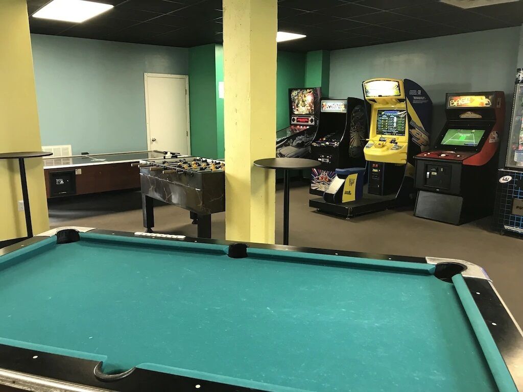 Un tavolo da biliardo, un tavolo da calcio e diverse macchine arcade in una stanza con pareti verdi e una porta sullo sfondo.