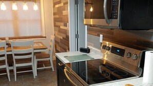Una cocina moderna con horno empotrado, microondas y estufa de acero inoxidable. Hay una tabla de cortar de madera en la encimera y un área de comedor con una mesa y sillas.