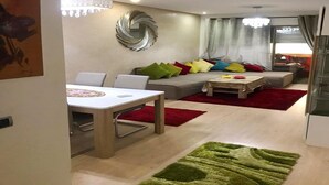 Een moderne woonkamer met een eethoek, een bank met kleurrijke kussens, een kroonluchter en een groot abstract kunstwerk aan de muur.