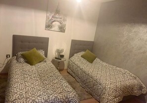 Een kamer met twee bedden, elk voorzien van een beddekleed met een patroon en een groen kussen. Tussen de bedden staat een klein tafeltje met een lamp en een decoratief object. Op de muur hangt een poster van de Eiffeltoren.