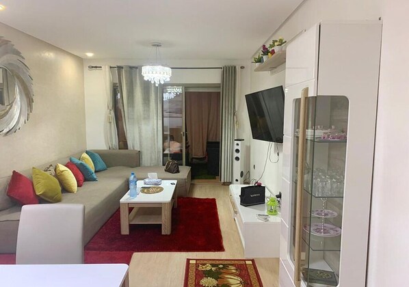 Een moderne woonkamer met een bank, salontafel en televisie. Er ligt een rood tapijt, een glazen kast met decoratieve objecten en er hangt een kroonluchter.