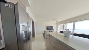 A modern kitchen with a stainless steel refrigerator, a flat-screen TV, and a large window overlooking the sea. - Modern Ocean View  Condo with Private  Beach   (PLAYAS encantada, playa de Rosarito)