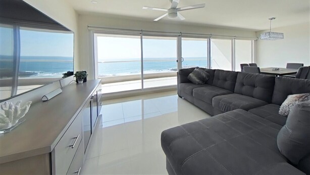A spacious living area with a large gray sofa, a dining table with chairs, and a ceiling fan. - Modern Ocean View  Condo with Private  Beach   (PLAYAS encantada, playa de Rosarito)