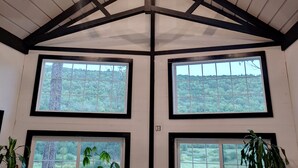 A room with a high ceiling, wooden beams, and large windows offering views of a green landscape. - Name: Hill Side<br>Country Setting. Heated Pool.  4 minutes to Dreams Park. (Cooperstown)