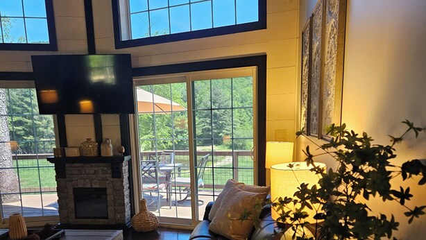 A spacious living room with a fireplace, a large window, and a view of the outdoors. - Name: Hill Side<br>Country Setting. Heated Pool.  4 minutes to Dreams Park. (Cooperstown)