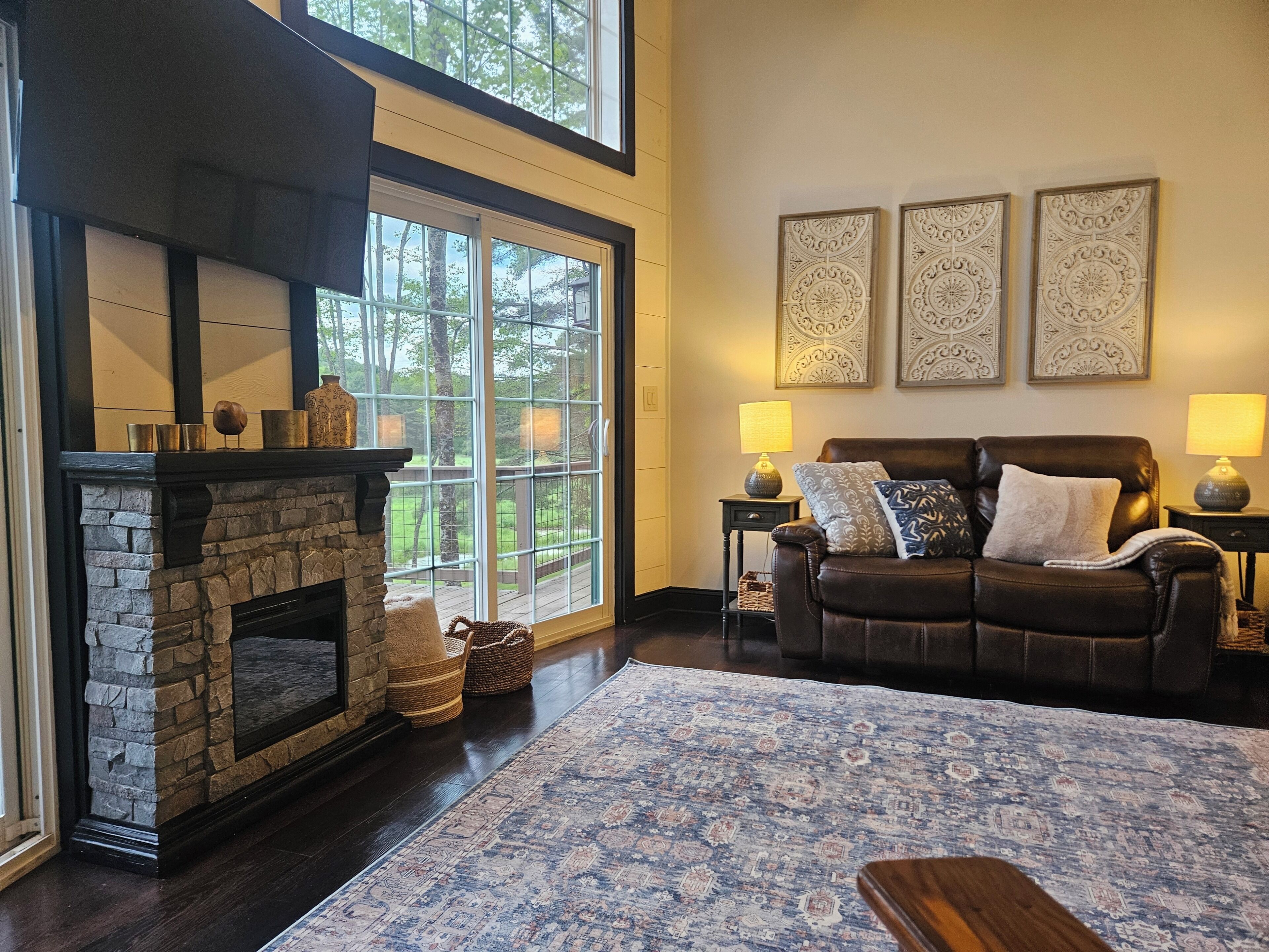 A living room with a stone fireplace, a leather sofa, a patterned rug, and large windows with a view of trees.