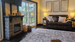 A living room with a stone fireplace, a leather sofa, a patterned rug, and large windows with a view of trees. - Name: Hill Side<br>Country Setting. Heated Pool.  4 minutes to Dreams Park. (Cooperstown)