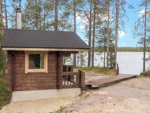 Una cabaña pequeña con fachada de madera, una chimenea y un muelle que se extiende sobre un lago rodeado de árboles.
