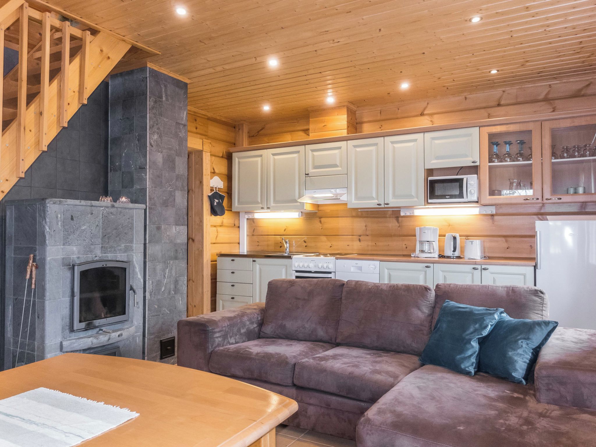 Un interno accogliente di una baita con camino in pietra, soffitto in legno e angolo cottura con mobili bianchi.