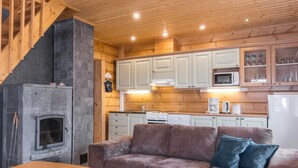 Interior acolhedor de uma cabana com lareira de pedra, teto de madeira e uma kitchenette com armários e eletrodomésticos brancos.