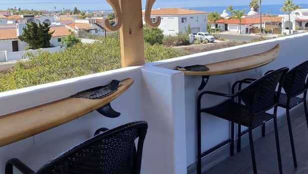 A wooden pergola with string lights, a surfboard, and black wicker chairs overlooking a coastal town. - Casa Tortugas - a Cozy, Relaxing House in a Gated Oceanfront Resort (Ensenada)