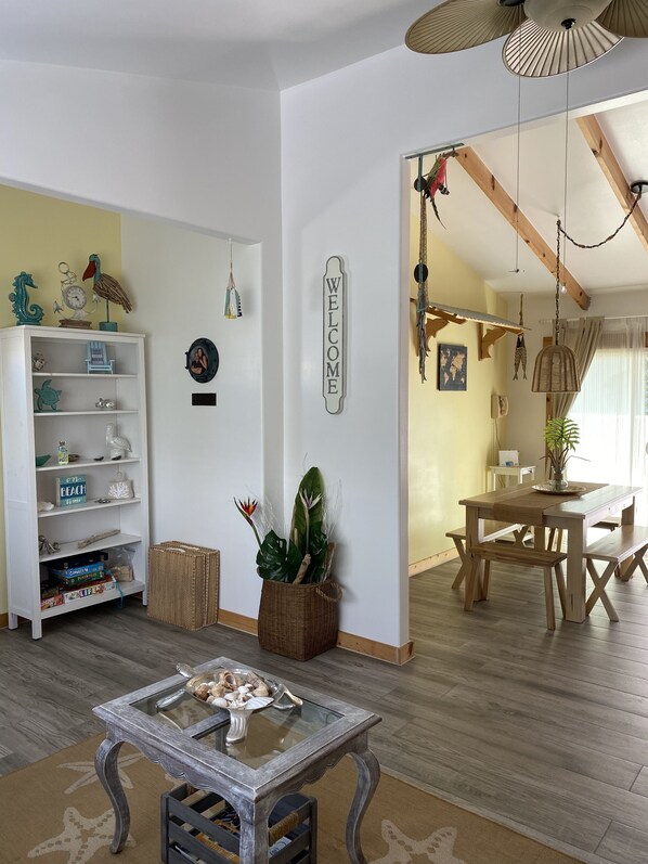 Une pièce comprenant une table à manger en bois, une table basse avec un plateau en verre, une bibliothèque blanche et un panneau « BIENVENUE » accroché au mur.