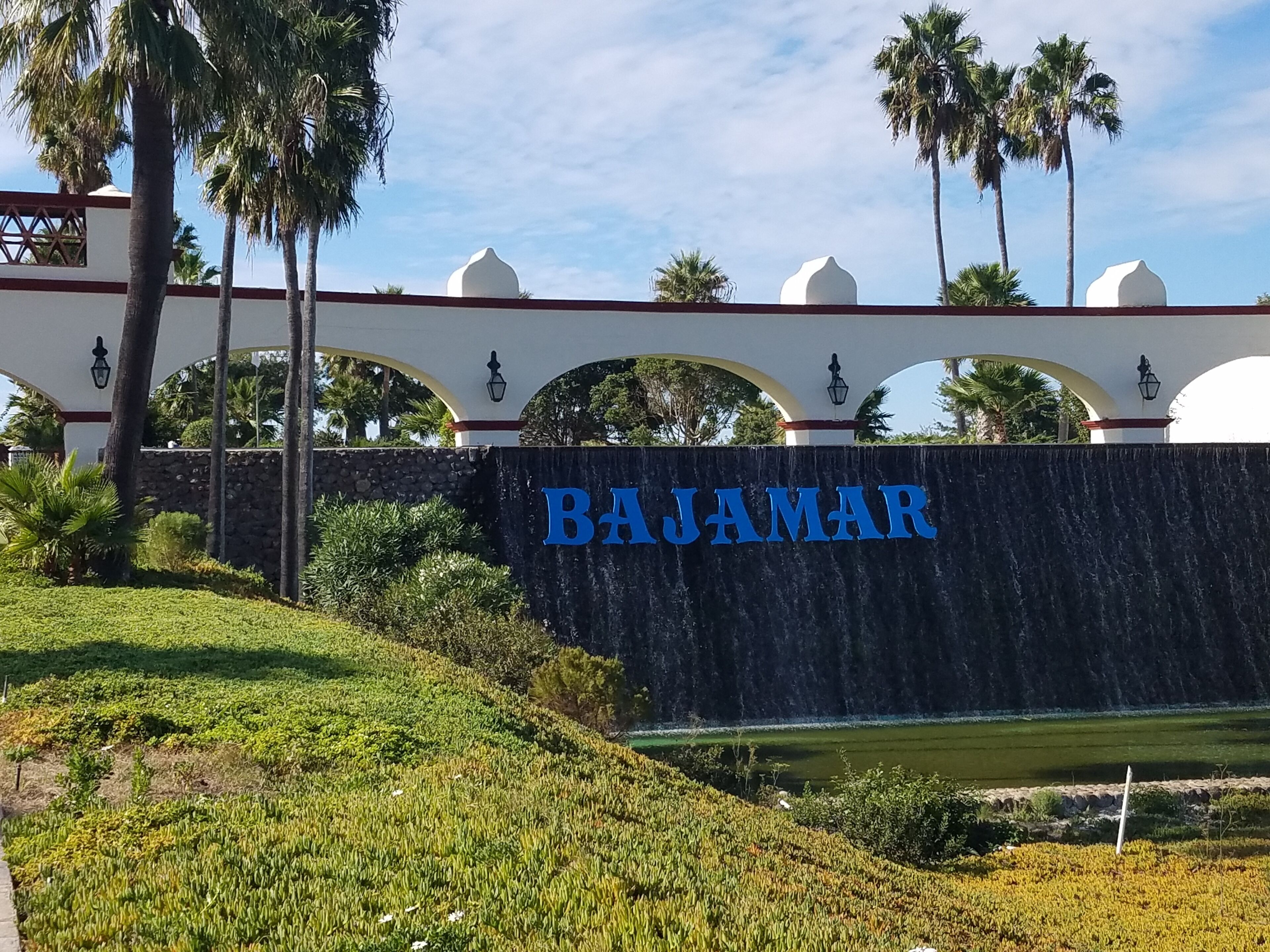 A sign with "BAJAMAR" in blue letters on a stone wall, surrounded by greenery and palm trees.