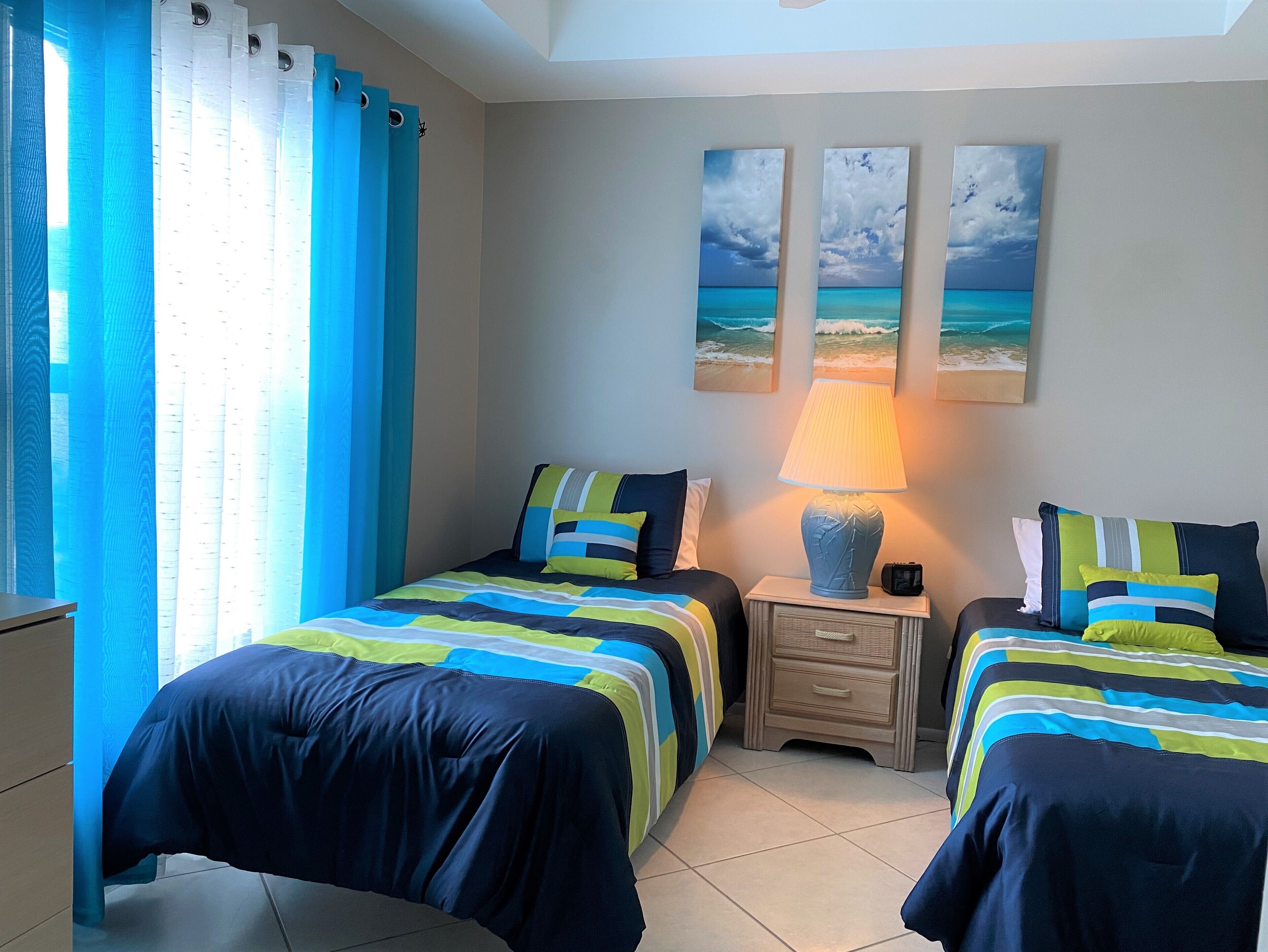 A room with two beds, a nightstand, and a lamp. There are three paintings on the wall depicting beach scenes.