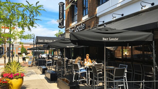 Un espace extérieur aménagé avec des tables et des chaises, une enseigne indiquant « Bar Louie », et un grand bac à fleurs.