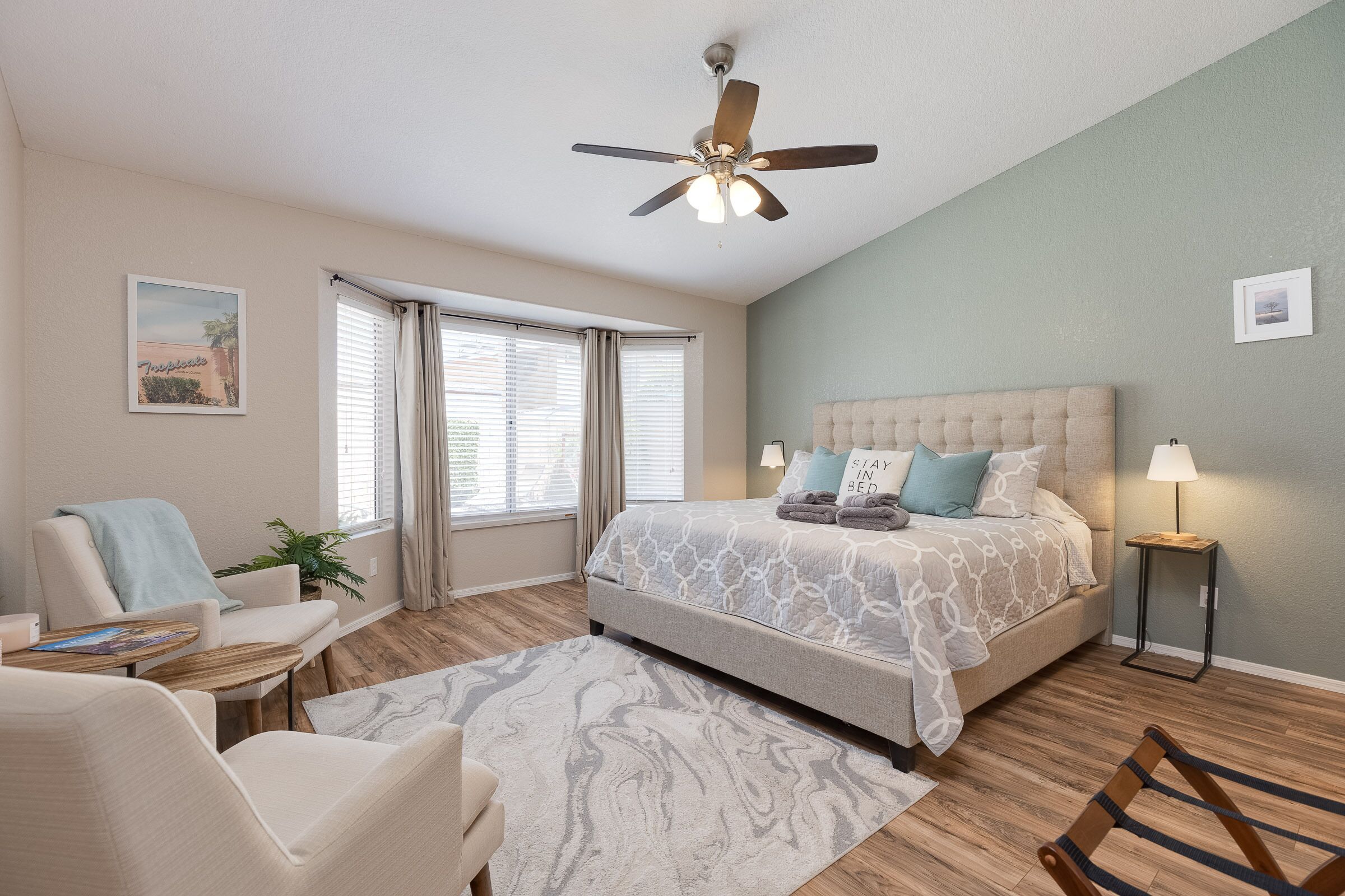 A bedroom with a bed, a chair, a table, a ceiling fan, and a window with curtains.