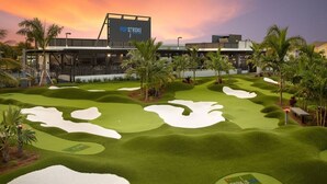 Un parcours de golf avec des bunkers de sable et des palmiers, un panneau indiquant « POF STROKE », et un bâtiment en arrière-plan.