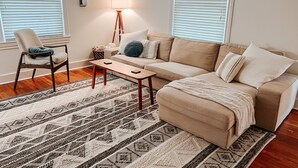 Ein Wohnzimmer mit einem beigen Sofa, einem Holztisch, einem gemusterten Teppich und einer Stehlampe.