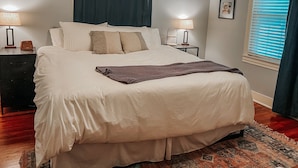 Ein ordentlich bezogenes Bett mit weißer Decke und Kissen, ein Nachttisch mit Lampe, eine kleine Ablagefläche und ein Fenster mit Jalousien.