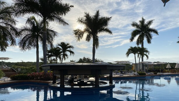A pool with a central gazebo, surrounded by palm trees and lounge chairs, with a view of mountains in the background. - Vidanta Luxury Paradise by (Nuevo Vallarta)