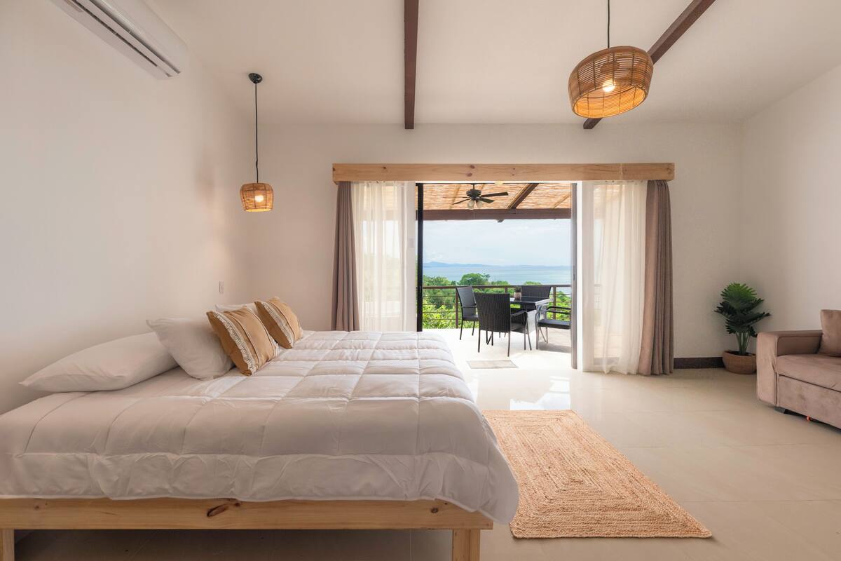 A bedroom with a large bed, a wooden bench, a woven light fixture, a ceiling fan, and a view of the outdoors through a sliding door.