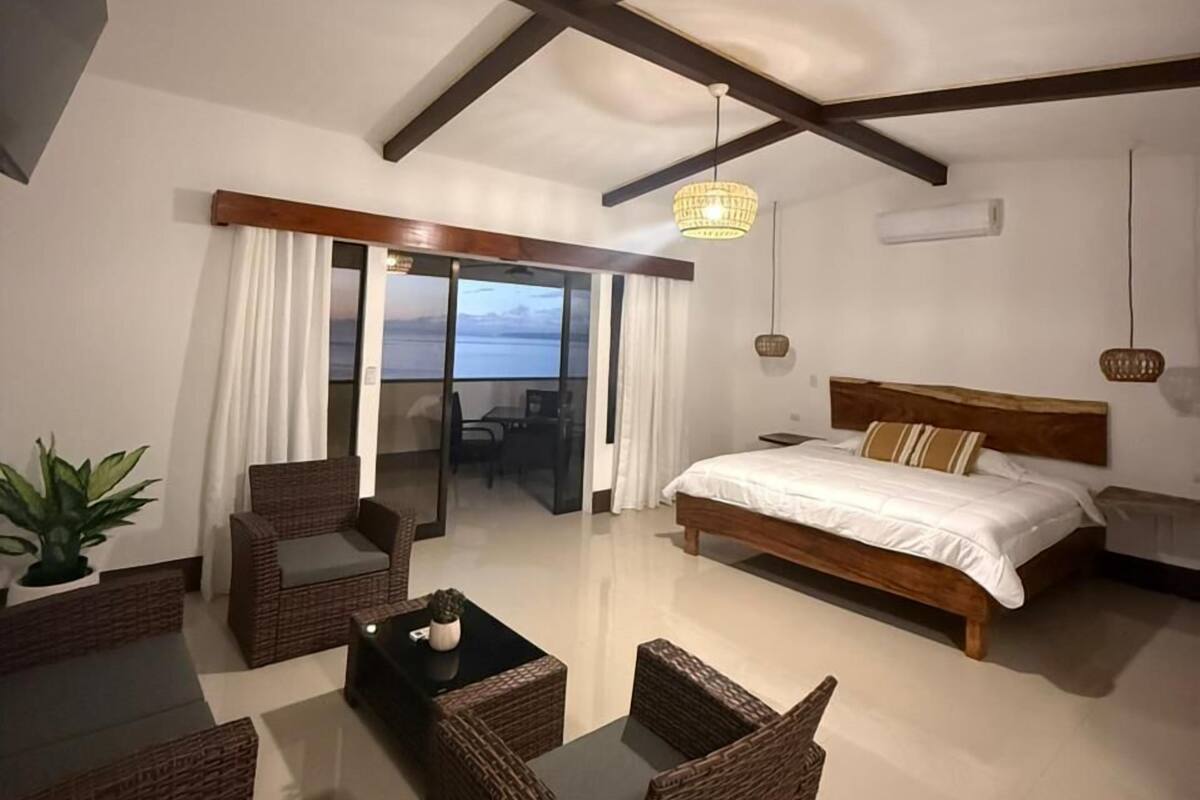 A bedroom with a large bed, a balcony with curtains, wicker furniture, and a ceiling with exposed beams.