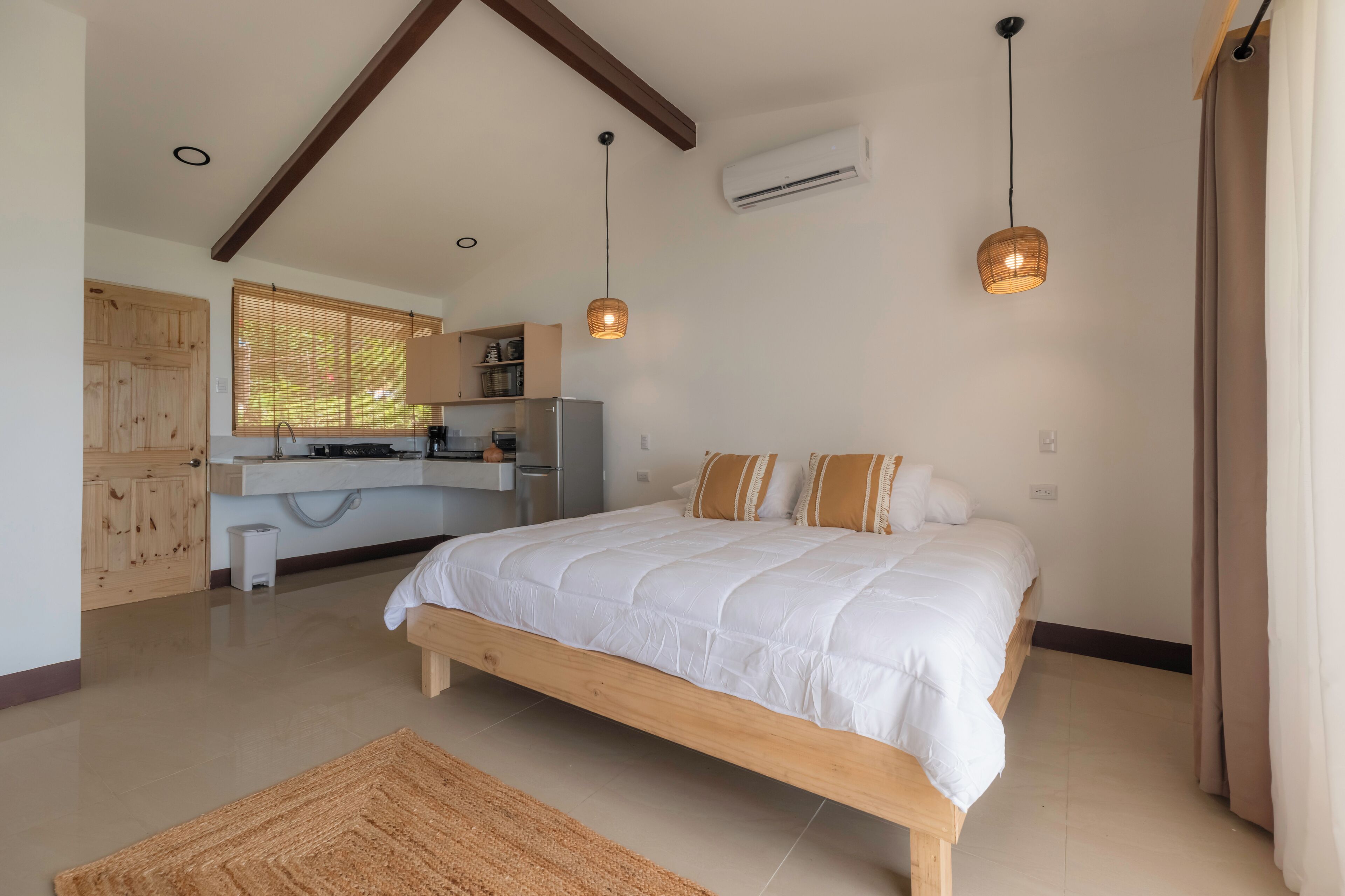 A bedroom with a wooden bed, a kitchen area with a refrigerator, and a window with blinds.