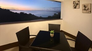 Un espace repas comprenant une table et des chaises, un ventilateur de plafond et une grande fenêtre offrant une vue sur un paysage au coucher du soleil.