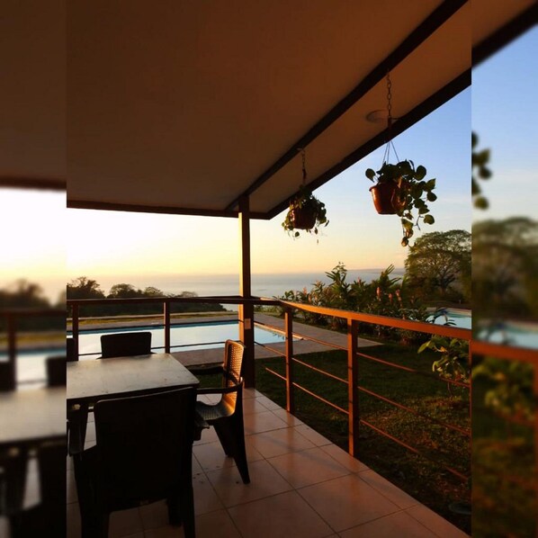 A covered outdoor area with a pool, a table, and chairs, overlooking a landscape with trees and a body of water. - Villas Azul (Tárcoles)