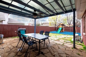 A covered patio with a stone floor, a table with chairs, and a grill.