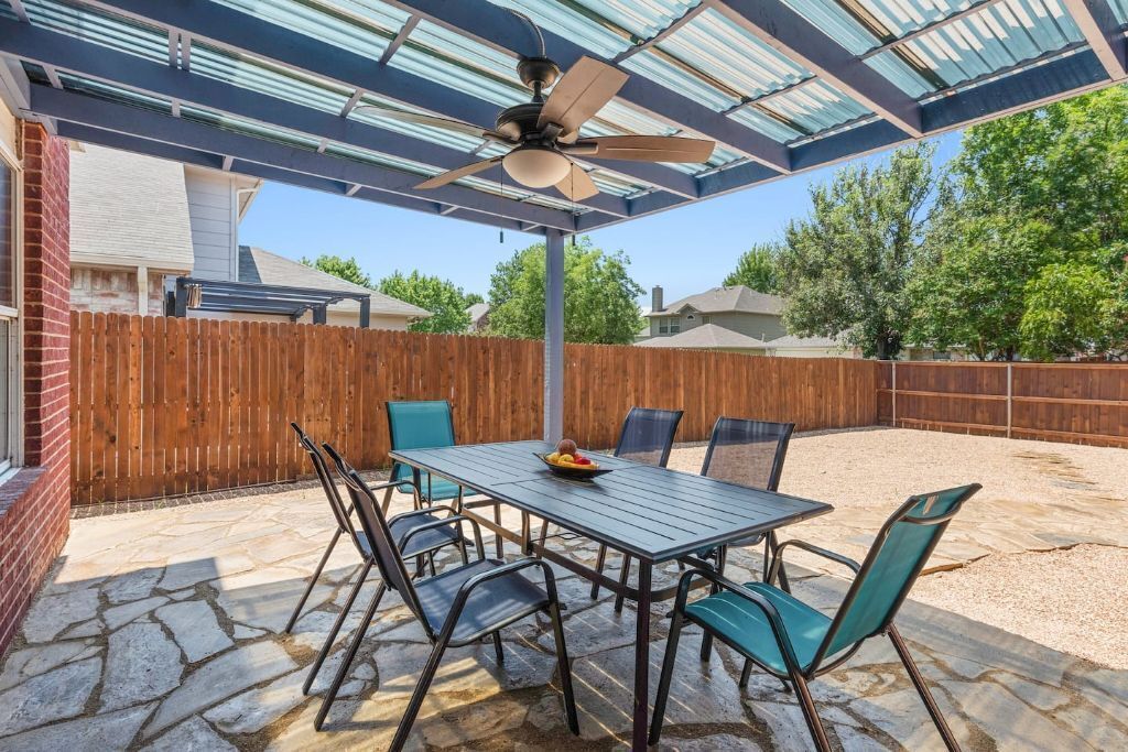 Une terrasse couverte avec une table et des chaises, un ventilateur de plafond et une clôture en bois en arrière-plan.