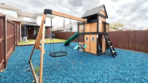 A wooden playhouse with a slide and swing set in a backyard with blue pebbles. - Stylish House w playset ,Game room &BBQ near DFW airport ,Gaylord sleeps 10-14 (Flower Mound)