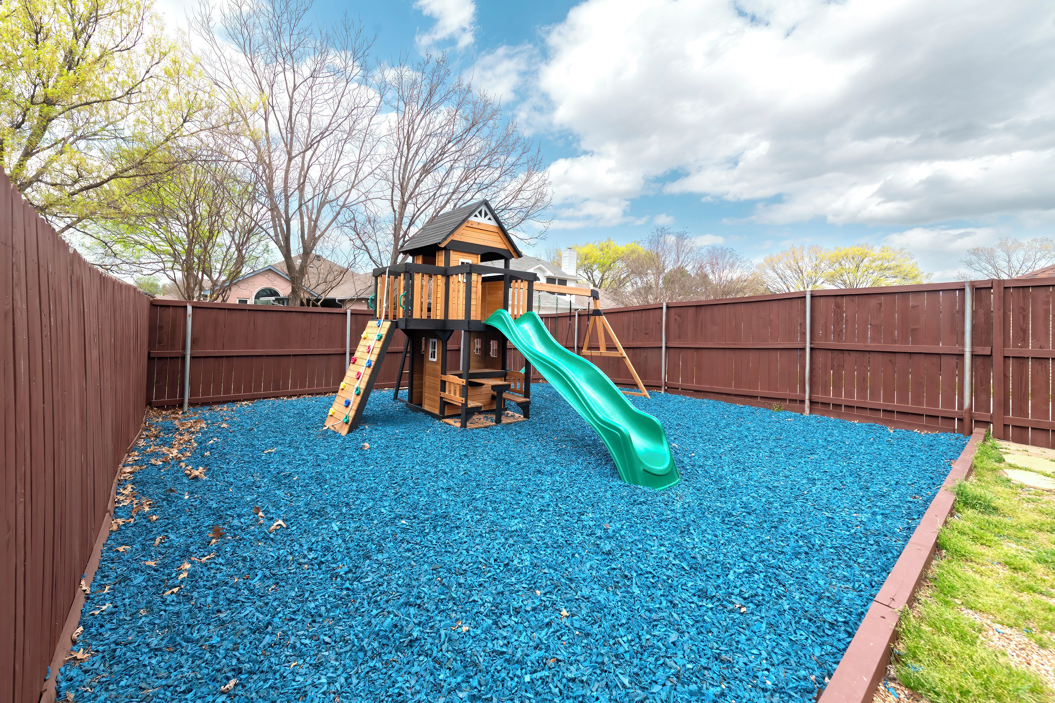 A wooden play structure with a slide and climbing wall, surrounded by a fence and blue pebbles.