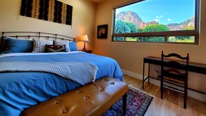 Un dormitorio con una cama grande, una silla de madera, una mesita de noche con una lámpara y una ventana con vistas a la montaña.