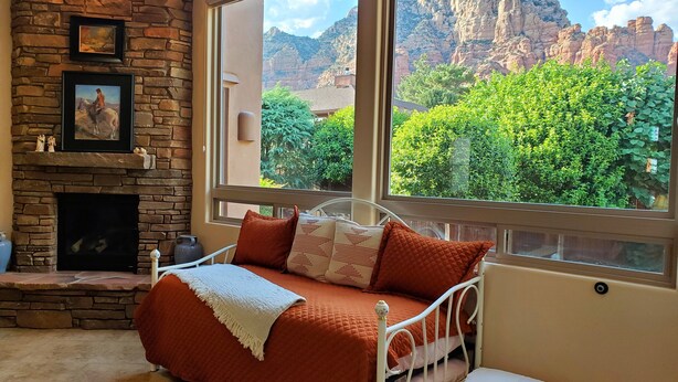 Un dormitorio con chimenea de piedra, una cama con manta roja, una ventana con vistas a la montaña y una mesita con objetos decorativos.