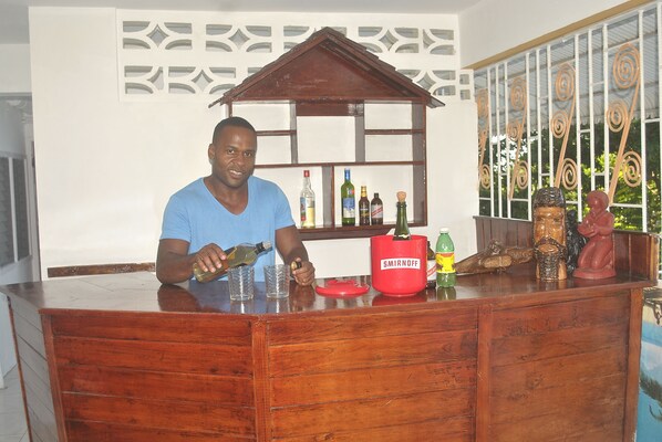 A man pouring a drink at a wooden bar counter with a shelf behind him holding various bottles. - Superb Villa in Montego Bay (Montego Bay)