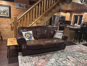 A wooden cabin interior with a leather sofa, a wooden table, a patterned rug, and a kitchen area with modern appliances.
