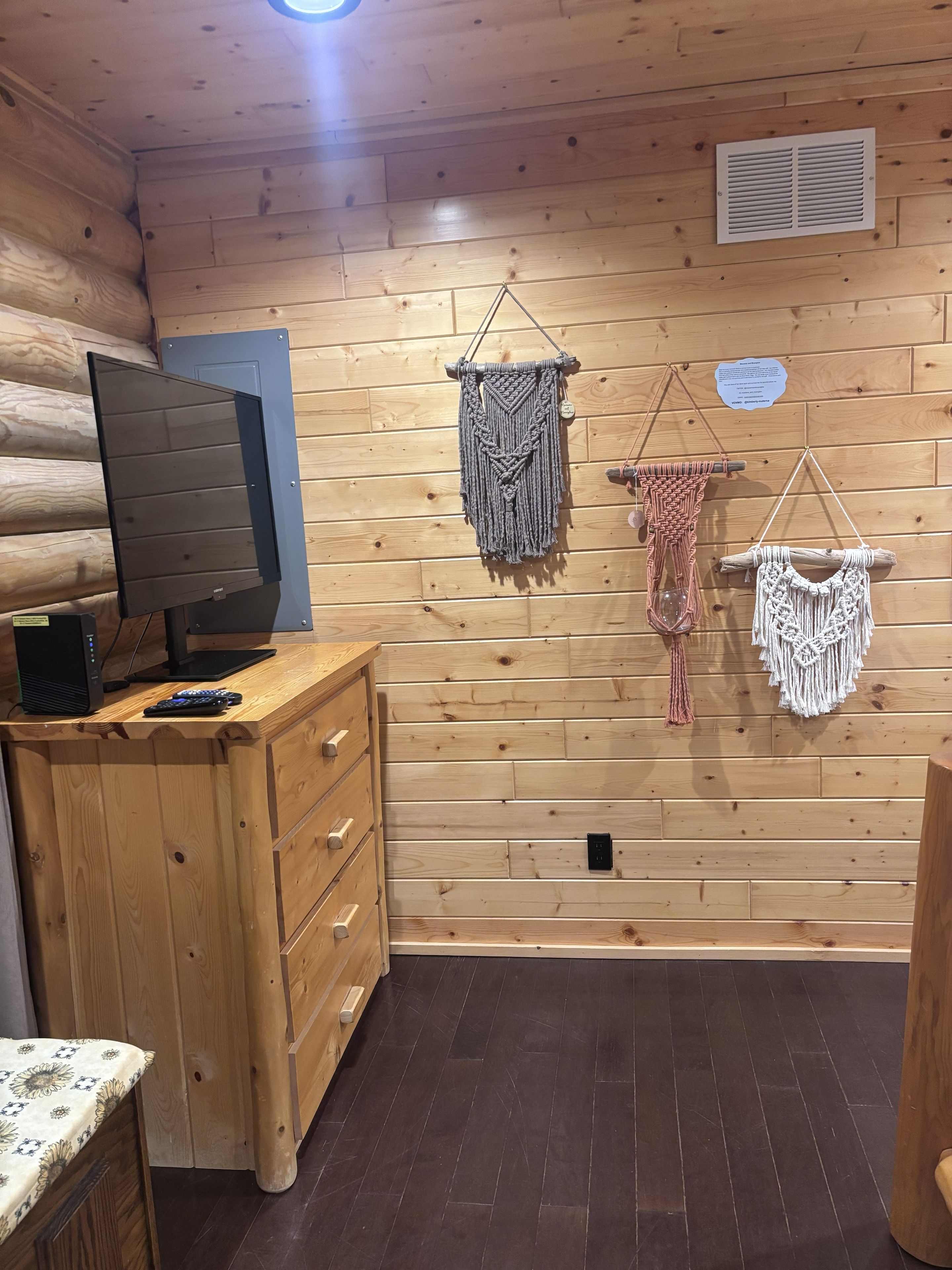 A wooden cabin interior with a television on a wooden dresser, a wall-mounted air vent, and three macramé wall hangings.