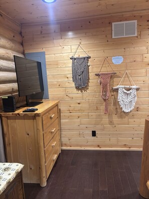 L’intérieur d’un chalet en bois, avec une télévision posée sur une commode en bois, une bouche d’aération fixée au mur et trois décorations murales en macramé.