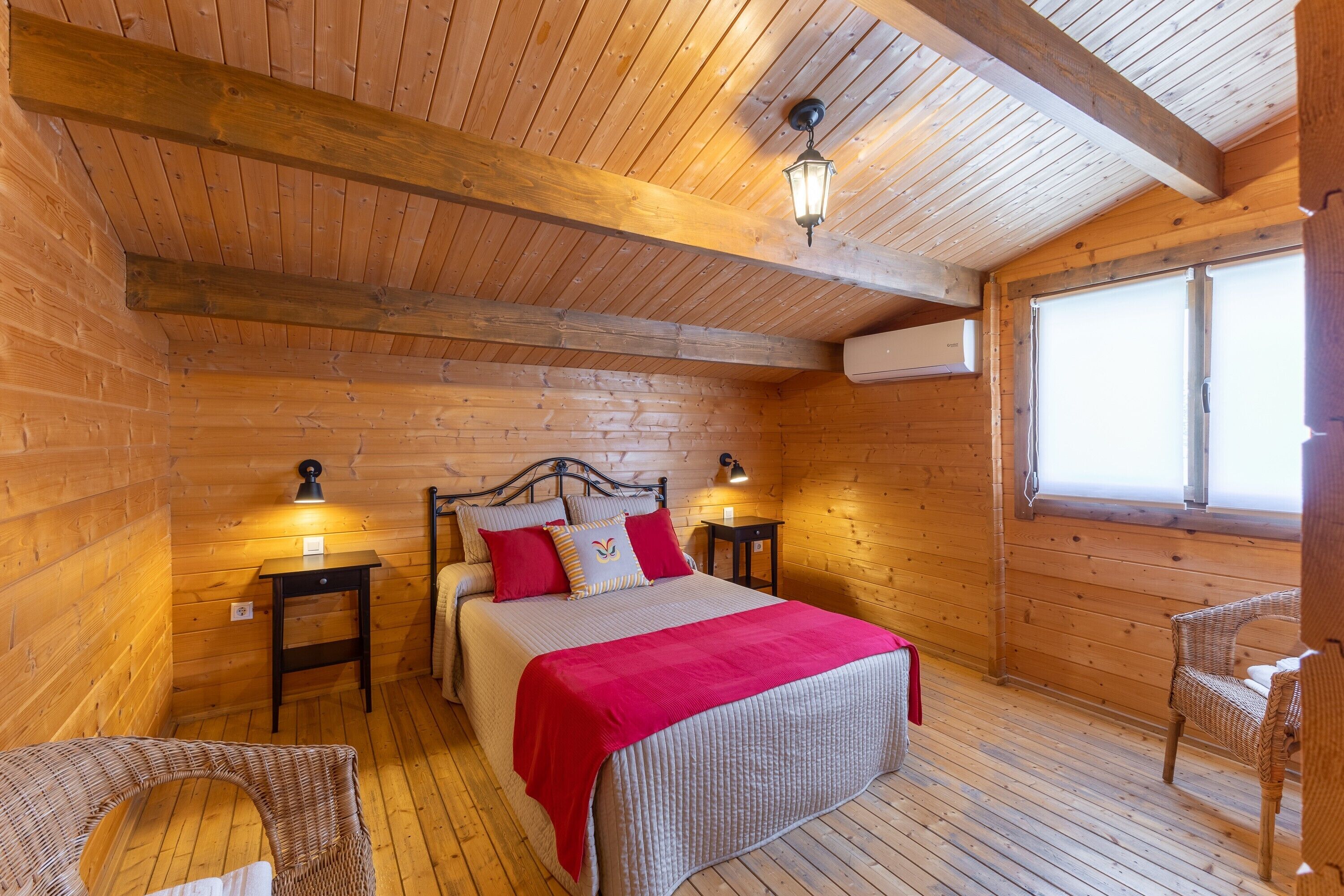 Un dormitorio de cabaña de madera con una cama, dos mesitas de noche, una manta roja, una silla de mimbre y una ventana.