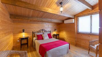 Un dormitorio de cabaña de madera con una cama, dos mesitas de noche, una manta roja, una silla de mimbre y una ventana.