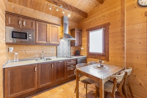 A wooden cabin interior with a kitchen featuring wooden cabinets, a microwave, a sink, and a dining table with chairs. - Casa Rural 'La Dehesilla' in Toledo – Cozy Retreat for 4 with Private Plot (Cobisa)