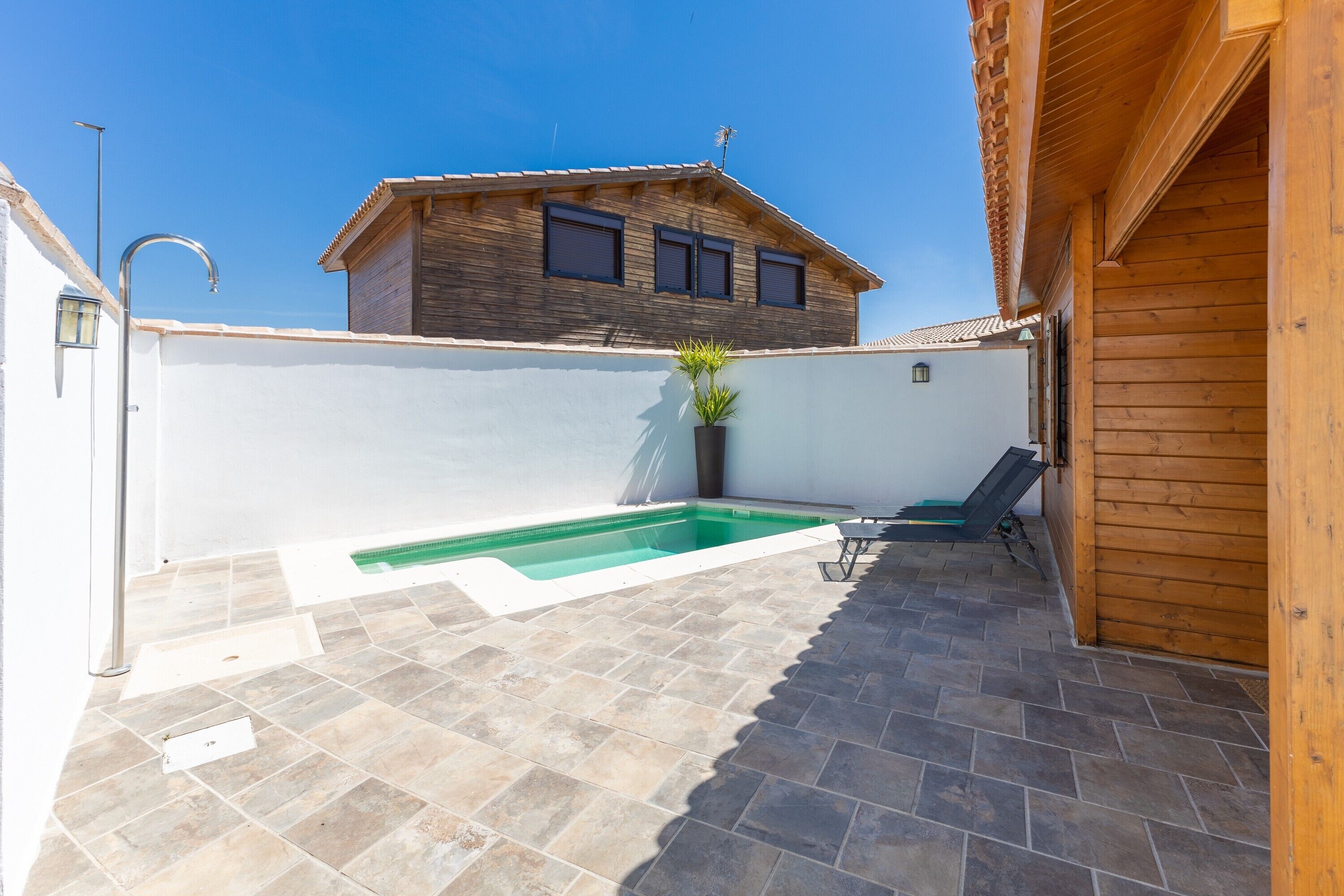 A rooftop patio with a swimming pool, a wooden house in the background, and a clear blue sky.